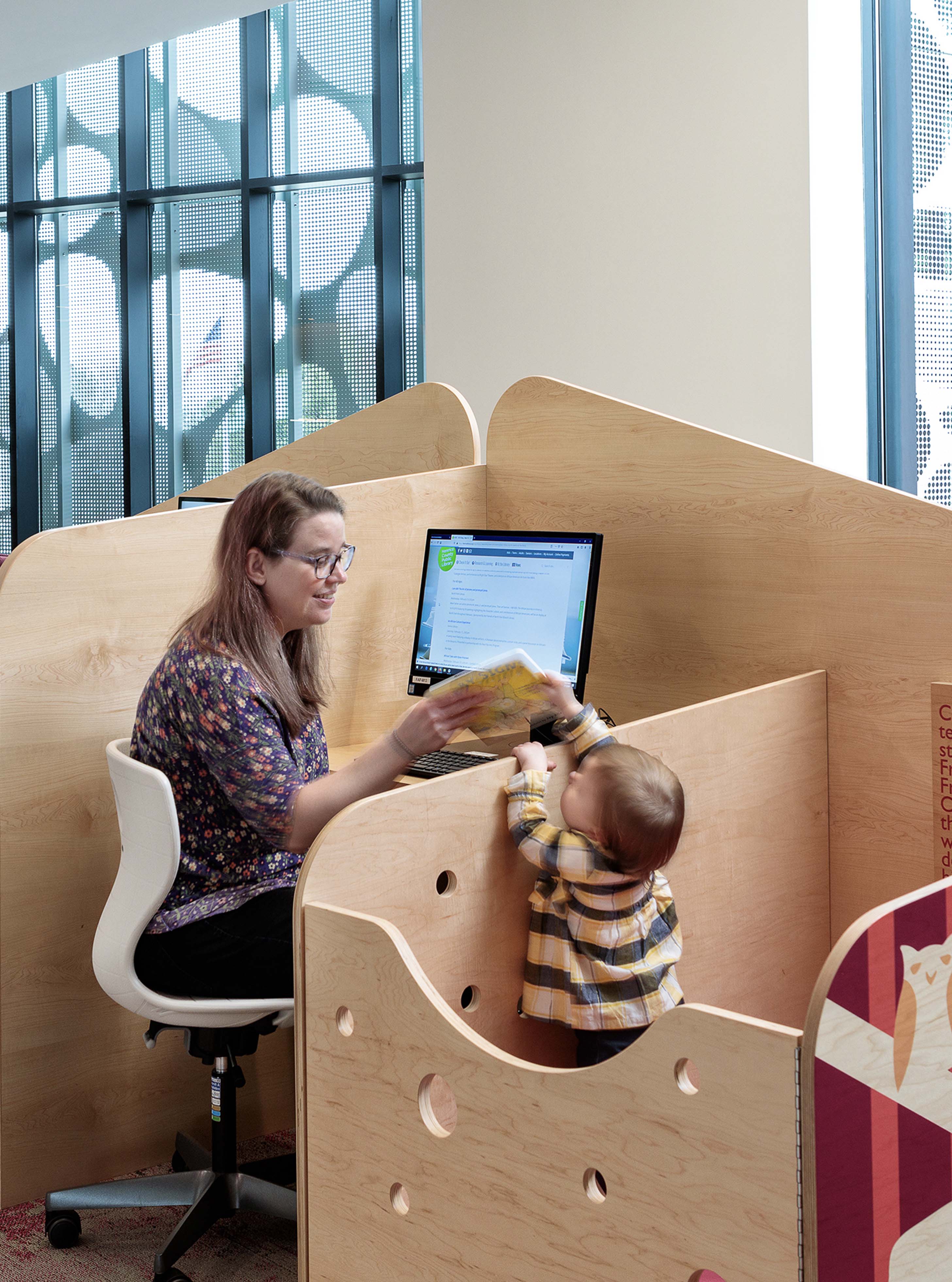 A child playing in playpen as a woman sits at attached desk