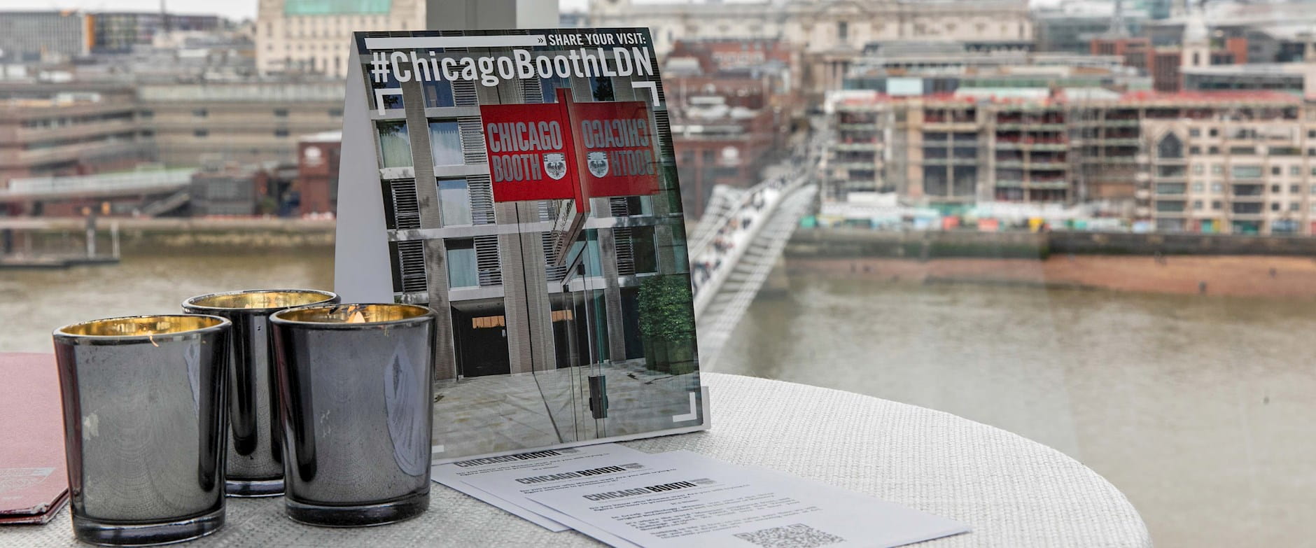 A placard sits next to 3 candles that reads "Share your visit: #ChicagoBoothLDN"; in the background is the River Thames